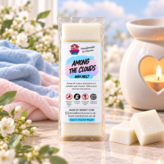 A packaged wax melt labeled 'Among the Clouds Wax Melt' stands on a reflective surface with two unwrapped square wax melts in front and a lit candle in a cream ceramic holder blurred in the background, alongside soft pink and blue fabric and white flowers.
