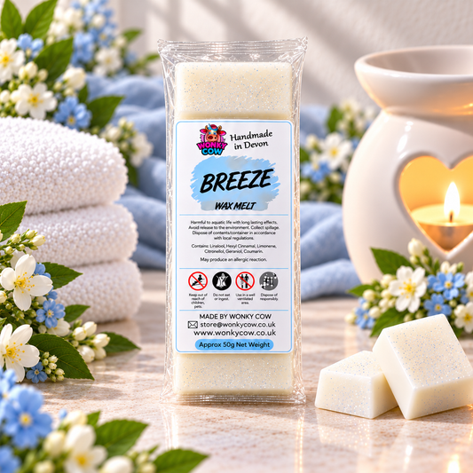 A package of Wonky Cow Breeze Wax Melts stands on a marble surface with two unwrapped off-white wax melts in front. The scene is styled with folded white towels, blue and white flowers, green leaves, and a lit white candle in a ceramic holder.