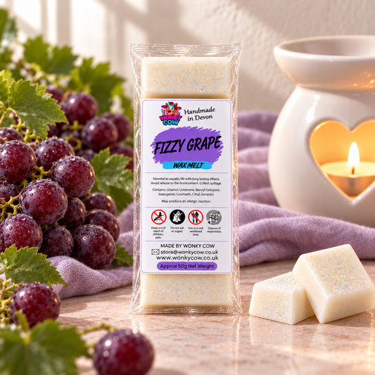 A clear plastic bag of Worksy Cow 'Fizzy Grape' wax melts stands on a marble surface, surrounded by red grapes, a lit heart-shaped candle holder, and two white wax melts with glitter flakes.