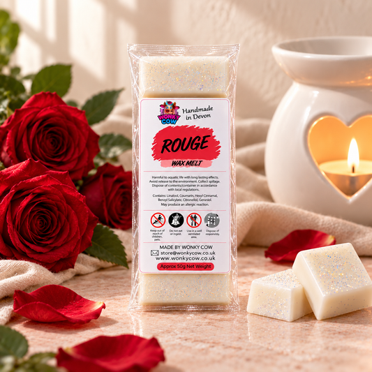 A packaged wax melt labeled 'Rouge Wax Melt' by Wonky Con, standing upright in a clear plastic sleeve. The product is surrounded by red roses, rose petals, and two pale glittery wax bars on a beige surface, with a white heart-shaped candle holder and lit candle in the background.