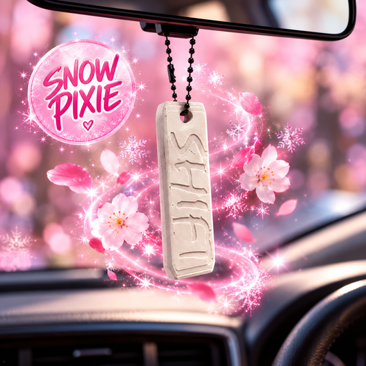 A car air freshener or hanging ornament with the word 'SNOW' carved into a rectangular beige tag, suspended from a black bead chain near the rearview mirror. The scene is decorated with pink cherry blossoms, petals, sparkles, and a pink circular badge reading 'SNOW PIXIE'.