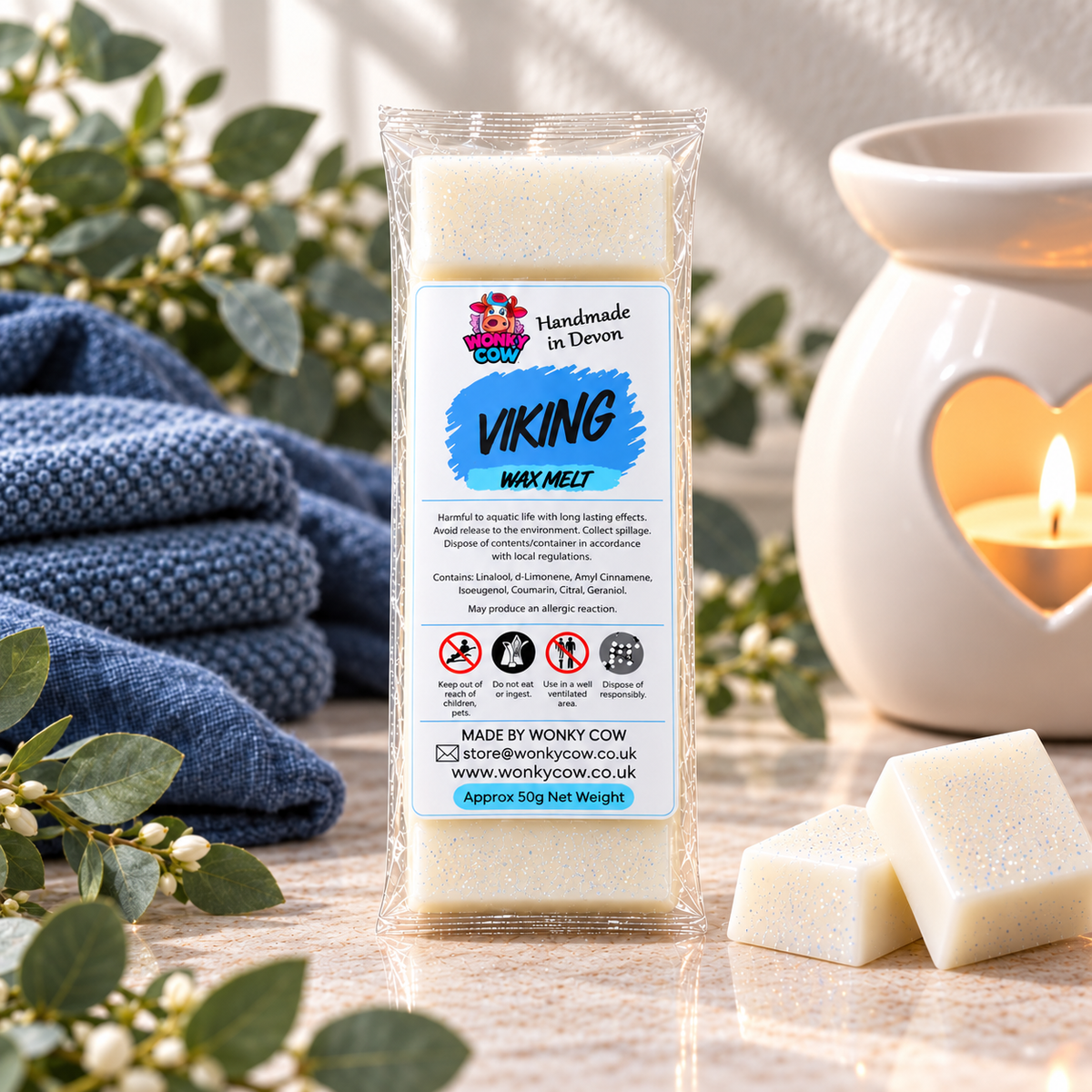 A packaged wax melt labeled 'Viking' by Wonky Cow, standing on a tabletop with a blue knit blanket, greenery, and a lit heart-shaped candle in the background.